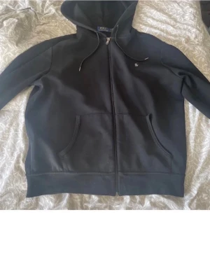 Svart hoodie från Polo Ralph Lauren - Svart hoodie med dragkedja från Polo Ralph Lauren. Klassisk design med huva, dragsnören och två fickor framtill. Liten broderad logga på bröstet. Perfekt för en chill och stilren look. Priset kan diskuteras vid snabb affär 🙏
