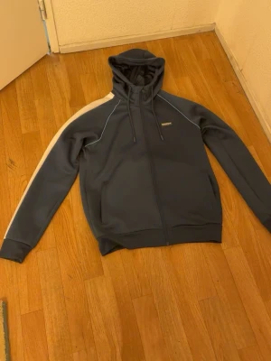 Blå hoodie med vit rand från McKenzie - Snygg blå hoodie från McKenzie med dragkedja och huva. Har en vit rand längs ena ärmen och diskret logga på bröstet. Tillverkad i mjukt och sportigt material, perfekt för en avslappnad stil. Fickor på sidorna och justerbar dragsko i huvan.