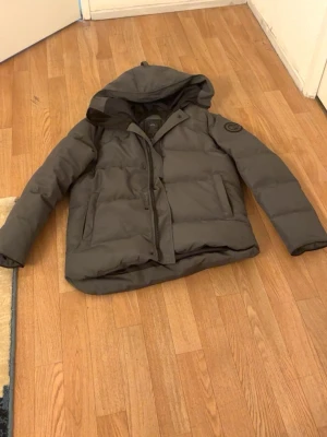 Mörkgrå pufferjacka med huva - Säljer en mörkgrå pufferjacka från Canada Goose med stor huva och diskret logotyp på ärmen. Jackan har två sidofickor, dragkedja och tryckknappar framtill. Perfekt för kalla vinterdagar och riktigt skön att bära.