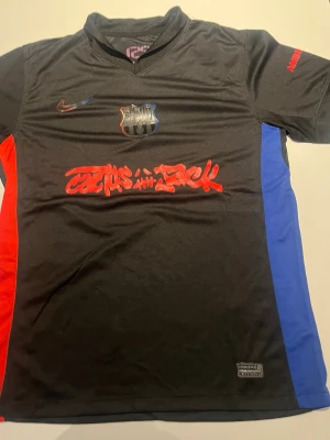 Svart FC Barcelona matchtröja Nike - Säljer en svart FC Barcelona matchtröja från Nike med klubbmärke och Nike-logga på bröstet. Tröjan har röda och blå sidopaneler, grafitti-tryck på framsidan och UNHCR-logga på ryggen. Tillverkad i lätt och ventilerande material, perfekt för fotboll eller träning.