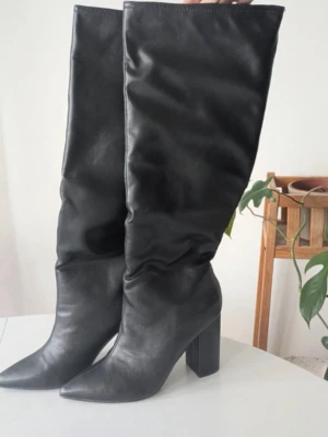Svarta höga boots med klack - Säljer ett par svarta höga stövlar i slätt skinnimitation med spetsig tå och bred blockklack. Stövlarna har en slouchy passform och når upp till knäet. Perfekta för dig som vill ha en edgy och trendig look. Från nelly och pris kan alltid diskuteras ska flytta och vill få bort då jag har för många liknande 