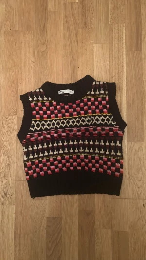 Mönstrad stickad väst från Zara - Säljer en stickad väst från Zara med färgglatt mönster i svart, rosa, rött, vitt och gult. Västen är ärmlös, har ribbad kant vid hals och nederkant samt en croppad passform. Perfekt för lager på lager och ger en retrovibe till din outfit.