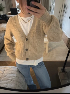 Beige stickad kofta med knappar - Mysig beige stickad kofta med v-ringning och tre mörka knappar framtill. Koftan har en avslappnad passform och ribbstickade muddar vid ärmslut och nederkant. Perfekt att slänga över en t-shirt för en chill look.