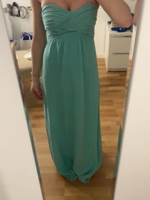 Ljusblå axelbandslös långklänning - Säljer en elegant ljusblå långklänning med draperad överdel och axelbandslös design. Klänningen har en vacker omlott-detalj upptill och ett flytande fall. Perfekt för bal eller lite finare tillfällen. Lite missfärgning på insidan men inget som förstör utsidan.