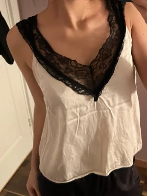 Vit body med svart spets - Snygg vit body med djup V-ringning och breda svarta spetsdetaljer längs urringningen och axelbanden. Från stockh lm 