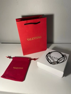 Valentino svart armband med logga - Stilrent svart armband från Valentino med justerbart band och silverfärgat metallspänne i form av Valentinos ikoniska V-logga. Kommer med originalförpackning och röd tygpåse med guldtext. Perfekt accessoar för en trendig look.