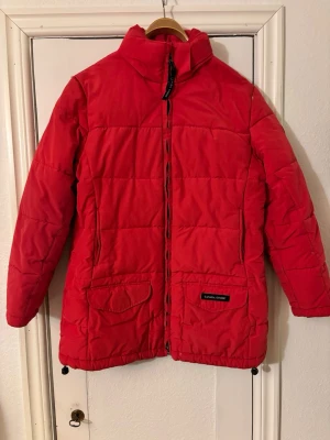 Röd dunjacka från Canada Goose - Snygg röd dunjacka från Canada Goose med hög krage, dragkedja framtill och två stora fickor med lock. Jackan har en klassisk quiltad design och är perfekt för kalla vinterdagar. Märkeslapp på ena fickan ger en exklusiv känsla.
