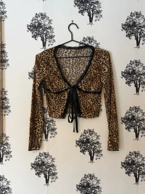 Leopardmönstrad croppad topp med knyt - Säljer en croppad leopardmönstrad topp med långa ärmar och djup v-ringning. Toppen har svarta spetsdetaljer längs urringningen och ett snyggt knyt framtill. Materialet är tunt och stretchigt, perfekt för att sticka ut.