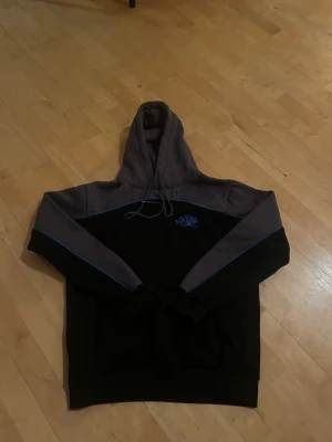 Svart och grå hoodie från Syna World - Snygg hoodie från Syna World i svart och grått med blå detaljer längs ärmarna och en liten blå logga på bröstet. Hoodien har huva med dragsko och är tillverkad i mjukt material, perfekt för en avslappnad streetwear-look.