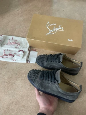 Loubs  - Snygga sneakers från Christian Louboutin i grå mocka med mörk grå nitar på tån och klassiska röda sulor. Modellen Louis Junior Spikes har rund tå, platt sula och svarta snören. Lyxig design med coola detaljer som sticker ut och ger en edgy vibe. Kvitto finns