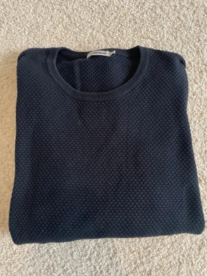 Mörkblå stickad tröja J.Lindeberg - Snygg mörkblå stickad tröja från J.Lindeberg med rund halsringning och diskret strukturmönster i tyget. Tröjan har ribbade muddar vid halsen och är tillverkad i ett mjukt material som känns skönt mot huden.