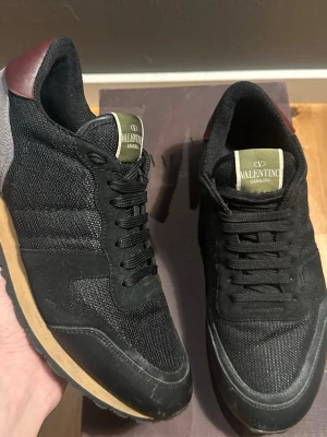 Valentino Garavani svarta sneakers - Snygga svarta sneakers från Valentino Garavani med detaljer i mocka och mesh. Skorna har en beige sula, vinröda detaljer vid hälen och en unik nitar-inspirerad design baktill. Perfekta för dig som vill ha en stilren och exklusiv look.