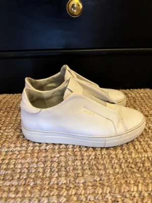 Vita sneakers från Axel Arigato - Stilrena vita sneakers från Axel Arigato i slät läder med minimalistisk design och guldfärgad logga på sidan. Skorna har låg profil och elastiska paneler istället för snörning för en clean look. Perfekta för dig som gillar enkel och modern stil.