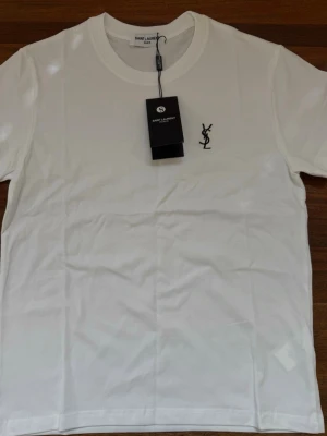 Vit Saint Laurent t-shirt med logga - Snygg vit t-shirt från Saint Laurent med klassisk svart YSL-logga broderad på bröstet. T-shirten har rund halsringning och korta ärmar. Tillverkad i mjuk bomull för en bekväm känsla och stilren look.
