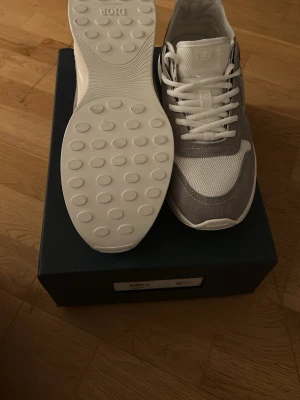 Dior gråvita sneakers i mesh och mocka - Snygga sneakers från Dior i grått och vitt med ovandel i mesh och mocka. Skorna har vita snören, diskret Dior-logga på plösen och en chunky vit sula med mönstrad undersida. Perfekta för dig som gillar stilrena och exklusiva sneakers.