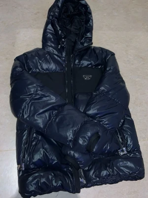 Svart pufferjacka från Alessandro - Snygg svart pufferjacka från Alessandro med glansig finish, huva och dragkedjor på sidorna. Jackan har en svart panel över bröstet och ryggen samt logga framtill. Perfekt för kalla vinterdagar och riktigt trendig stil.