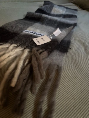 Rutig halsduk från Acne Studios - Snygg rutig halsduk från Acne Studios i mjuk ullmix. Färgskalan går i svart, grått och vitt med stora rutor och fransar i ändarna. Perfekt accessoar för att lyfta din outfit med en klassisk men modern touch.