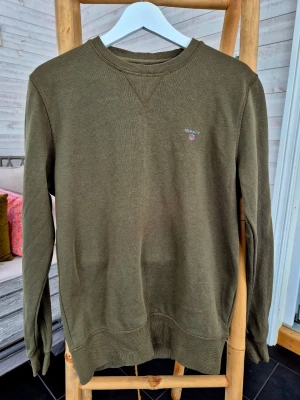 Olivgrön sweatshirt från Gant, storlek M - Olivgrön sweatshirt från Gant med rund halsringning och broderad logotyp på bröstet. Tröjan har ribbade muddar vid ärmslut och nederkant samt klassisk relaxed passform. 