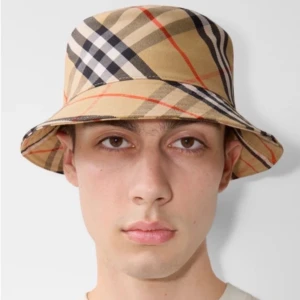 Burberry hatt  - Stilren bucket hat i klassiskt beige med Burberrys ikoniska rutiga mönster i svart, vitt och rött. Hatten har mjuk form och bred brätte, perfekt för en trendig och avslappnad look. Tillverkad i bomull.
