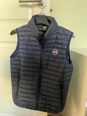 Mörkblå dunväst från Colmar - Snygg mörkblå dunväst från Colmar med quiltad design och hög krage. Västen har dragkedja framtill och två praktiska fickor med dragkedja. Perfekt för lager-på-lager och har en stilren look med Colmar-logga på bröstet. Fint skick.