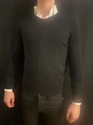 Svart v-ringad stickad tröja - Svart v-ringad stickad tröja med lång ärm och ribbade muddar vid ärmslut och nederkant. Enkel och stilren design som funkar till skjorta under. Passformen är normal och materialet känns mjukt och skönt mot huden.