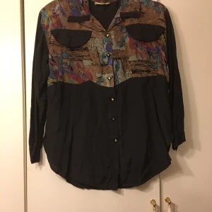 Mönstrad blus med knappar storlek S/M - Snygg blus med färgglatt mönster upptill och svart nedtill. Blusen har långa ärmar och knappar framtill. Perfekt för en unik stil.