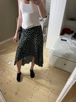 Blommig omlottkjol från Lindex i storlek S - Supersöt svart omlottkjol från Lindex med små vita och gula blommor. Kjolen är lång, har knyt i midjan och är tillverkad i mjuk polyester. Perfekt för en varm sommardag! Modellen är 172cm lång.