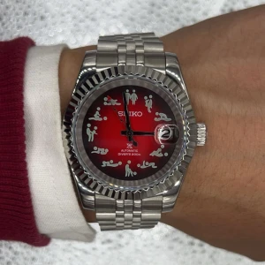 Seiko automatisk dykarklocka röd/silver - Unik Seiko automatisk dykarklocka med röd urtavla och roliga figurer som timmarkeringar. Boett och länk i silverfärgat stål, tydlig datumvisning och robust design. Urtavlan har en cool gradient från rött till svart. 