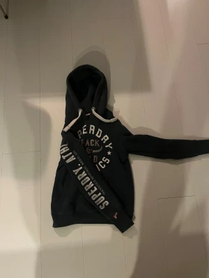 Mörkblå Superdry hoodie med tryck - Snygg mörkblå hoodie från Superdry med stort vitt tryck på bröstet och ärmen. Tröjan har huva med vita snören och en klassisk känguruficka framtill. Perfekt för dig som gillar sportig streetstyle och vill ha något bekvämt och coolt.
