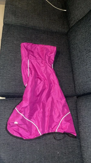 Lila regnjacka från Rukka, storlek 50 - Säljer en lila regnjacka från Rukka i storlek 50. Jackan är tillverkad i 100% polyester och har reflexdetaljer längs kanterna. Perfekt för regniga dagar och har en sportig vibe med smidig passform och svarta kantband.
