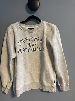 Grå sweatshirt Peak Performance 160 - Grå sweatshirt från Peak Performance med broderad text 'Sportswear Peak Performance' på bröstet. Tröjan har rund hals, ribbade muddar vid ärmslut och nederkant samt lång ärm. Perfekt för chill dagar och sportiga outfits.