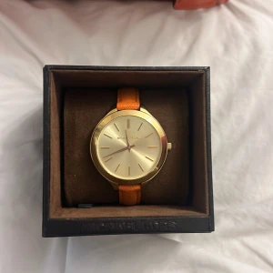 Guldig klocka från Michael Kors - Stilren och äkta klocka från Michael Kors med rund, guldig urtavla och orange armband i skinn. Klockan har minimalistisk design med tunna index och spänne i metall. Perfekt accessoar för dig som vill sticka ut med färg och klassisk look. Ordinarie pris: 2000kr. Passar bra som julklapp! Lite sliten i bandet, se bild.
