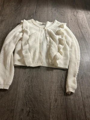 Vit stickad kofta med volanger Lindex - Supersöt vit stickad kofta från Lindex med volangdetaljer längs axlarna och framstycket. Koftan har rund hals, ribbade muddar och knappar framtill. Perfekt för dig som gillar en mjuk och cozy look med extra detaljer.