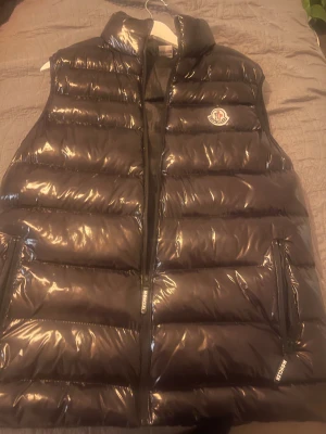 Svart dunväst från Moncler - Snygg svart dunväst från Moncler med glansig finish och klassisk logga på bröstet. Västen har hög krage, två dragkedjefickor och quiltad design. Perfekt för lager-på-lager och ger en cool streetstyle-vibe.