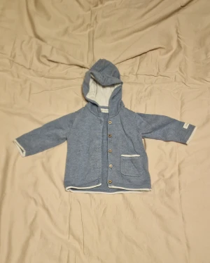 Blå finstickad tröja med knappar - Mysig blå stickad tröja med huva och träknappar framtill. Jackan har en liten ficka på vänster sida och vita detaljer längs kanterna. 