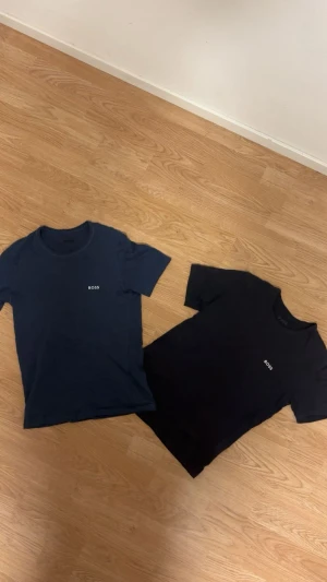 Svart och marinblå BOSS t-shirt - Två stilrena t-shirts från BOSS, en i svart och en i marinblå. Båda har rund hals, korta ärmar och en enkel BOSS-logga på bröstet. Materialet är mjuk bomull som känns skönt mot huden. Perfekt för dig som gillar clean och klassisk stil.