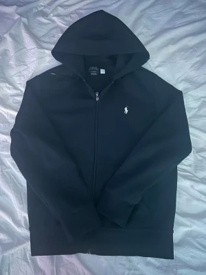 Svart hoodie från Polo Ralph Lauren - Svart hoodie från Polo Ralph Lauren med klassisk vit logga på bröstet. Tröjan har huva, hel dragkedja och långa ärmar. Materialet är mjuk bomull och polyester, perfekt för chill dagar. Snygg och enkel design som funkar till det mesta.