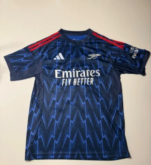 Arsenal bortatröja Gyökeres 14 Adidas - Arsenal bortatröja från Adidas med Gyökeres och nummer 14 på ryggen. Tröjan är mörkblå med blixtmönster i blått, röda axelränder och vita tryck. Tillverkad i lätt och ventilerande polyester, perfekt för match eller träning. Aldrig använd.