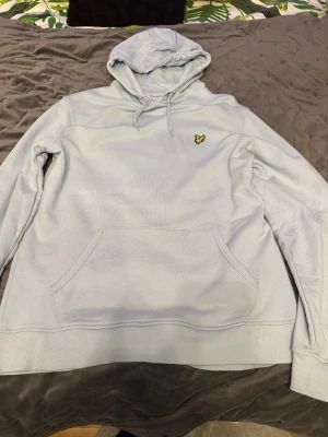 Ljusblå hoodie från Lyle & Scott - Snygg ljusblå hoodie från Lyle & Scott i ekologisk bomull. Klassisk design med huva och snörning, stor magficka och broderad gul fågel på bröstet. Perfekt för chill dagar och enkel att matcha med jeans eller joggers. Priset är ej hugget i sten.