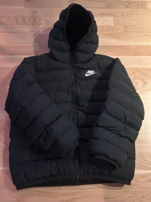 Svart pufferjacka från Nike med huva - Säljer en svart pufferjacka från Nike med huva och dragkedja framtill. Jackan har ett stilrent, quiltat mönster och Nike-logga på bröstet. Perfekt för kalla dagar och har en modern vibe! 