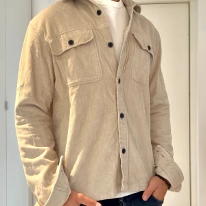 Beige manchester overshirt Lost Minds - Säljer en beige overshirt från Lost Minds i mjuk manchester. Skjortan har två stora bröstfickor med lock och svarta knappar framtill. Perfekt lager-på-lager-plagg med avslappnad passform och klassisk krage.