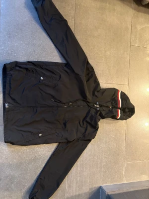 Svart Moncler vindjacka  - Svart vindjacka från Moncler med hel dragkedja, huva med röd och vit rand, samt två fickor framtill. Jackan har Moncler-logga på ärmen och är tillverkad i lätt polyester. Perfekt för dig som vill ha en clean och sportig look.