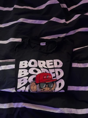 Svart Bored of Directors t-shirt - Svart t-shirt från Bored of Directors med stor tryckt apa i röd keps och solglasögon på bröstet. Texten 'BORED' upprepas i vitt bakom motivet. Rund hals och korta ärmar. Materialet är mjuk bomull, perfekt för dig som gillar streetwear och coola prints.