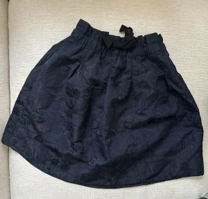 Blommig kortkjol från H&M - Supersöt kortkjol från H&M med blommigt mönster i tyget och en fin rosett framtill i midjan. Kjolen har en lätt puffig form och är gjord i polyester, vilket ger en snygg struktur och glans. 