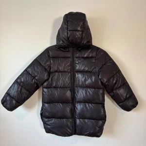Svart pufferjacka - 140 Storlek  - Svart pufferjacka från i storlek 140 med huva och dragkedja framtill. Jackan är quiltad är glansig. Klassisk modell med lång ärm och två sidfickor.