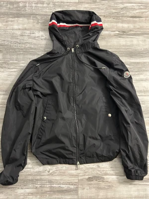 Svart vindjacka från Moncler med huva - Svart vindjacka från Moncler med huva och dragkedja framtill. Jackan har Moncler-logga på ärmen, snörning vid huvan och röda/vita detaljer på huvkanten. Tillverkad i lätt polyester, perfekt för blåsiga dagar. Klassisk och sportig look med två fickor framtill.