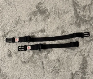 Svart justerbart bälte med spänne för ryggsäck - Säljer två svarta, justerbara bälten med plastspänne och liten röd logga på spännet. Bältena är smala och tillverkade i slitstark syntet med enkel design, perfekta för att hålla saker på plats eller som accessoar till väskor och utrustning.