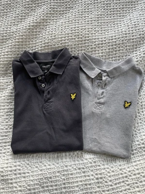 Två pikétröjor från Lyle & Scott - Säljer två klassiska pikétröjor från Lyle & Scott, en i mörkblå och en i ljusgrå. Båda har korta ärmar, krage och knappar framtill samt den ikoniska gula fågeln broderad på bröstet. Materialet är mjuk bomull som är skönt mot huden.