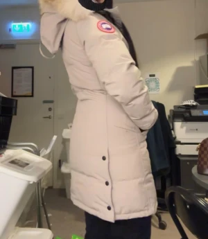 Beige vinterjacka från Canada Goose - Säljer en beige vinterjacka från Canada Goose med huva och pälskant. Jackan har svarta knappar, dragkedja och logotyp på ärmen. Perfekt för kalla dagar med sin varma fyllning och klassiska design. Väldigt unik från Johnells NYPRIS 13000