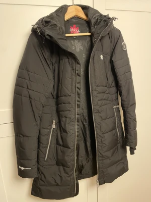 Svart lång dunjacka från Áhkká - Svart jacka  från Áhkká i fint skick. Tunnare modell av vinterjacka  Ena snöret har släppt från insidan, vid luvan. men inget som jag tänkt på. Se bilder  Inga andra revor eller märken
